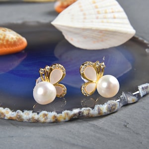 May include: A pair of gold butterfly earrings with white pearl accents. The earrings are set against a blue and white agate background.