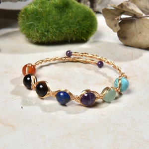 May include: A gold wire wrapped bracelet with seven colorful gemstone beads. The beads are orange, black, blue, purple, green, and turquoise.