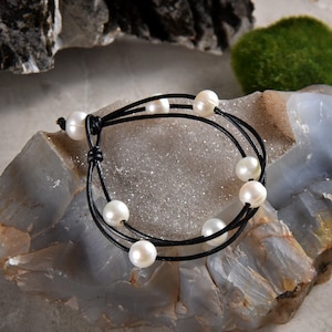 May include: A black leather bracelet with white pearls. The bracelet is made of three strands of leather and has seven pearls.