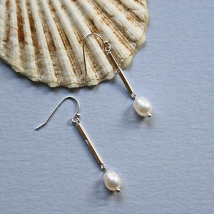 May include: A pair of silver dangle earrings with a small pearl hanging from each. The earrings are on a light blue background.