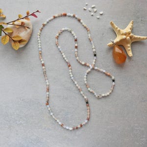 Puede incluir: Un collar largo de cuentas con un colgante en forma de lágrima. El collar presenta una variedad de pequeñas cuentas coloridas en tonos blancos, marrones y melocotón. El colgante es una piedra grande de color naranja. También hay una estrella de mar y elementos decorativos.