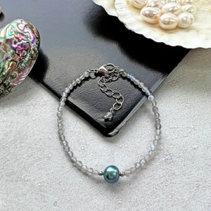 May include: A silver bracelet with a dark blue pearl charm and a silver clasp. The bracelet is made of small, round, light gray beads.