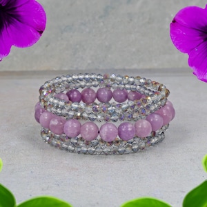 May include: Three stacked bracelets. The bracelets combine light purple, round beads with clear, faceted beads. The bracelets are arranged on top of each other. The background is a light grey colour with purple flowers.