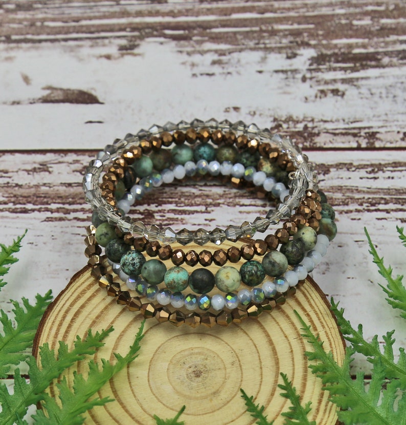 Puede incluir: Un conjunto de seis pulseras con una mezcla de cuentas turquesa y doradas. Las pulseras est&aacute;n apiladas sobre una superficie de madera.