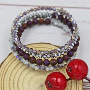 Puede incluir: Un conjunto de cuatro pulseras con un dise&ntilde;o en capas de cuentas moradas, plateadas y blancas. Las pulseras est&aacute;n apiladas sobre una superficie de madera.