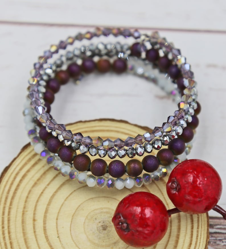 Puede incluir: Un conjunto de cuatro pulseras con un dise&ntilde;o en capas de cuentas moradas, plateadas y blancas. Las pulseras est&aacute;n apiladas sobre una superficie de madera.