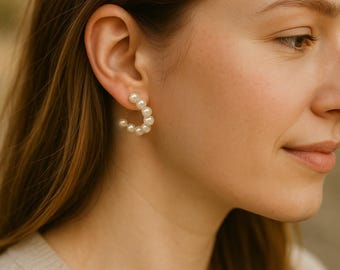May include: A close-up of a woman's ear adorned with a pearl hoop earring. The earring is a semi-circle design, composed of multiple small, round, white pearls. The woman has long, light brown hair and is wearing a light-colored sweater.