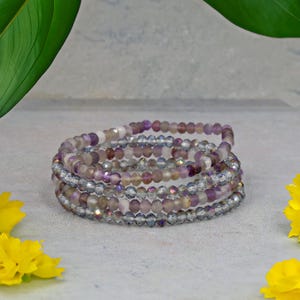 Puede incluir: Una pulsera envolvente de múltiples hilos con cuentas moradas, transparentes e iridiscentes. La pulsera está enrollada y descansa sobre una superficie gris claro. Flores amarillas y hojas verdes en el fondo.