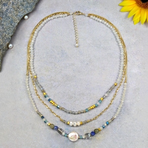May include: A multi-strand necklace with gold chains and beads. The top layer has small white beads and a gold chain. The middle layer features grey and blue beads. The bottom layer includes a large white pearl and various blue and grey beads.