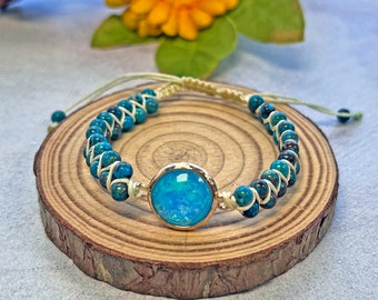 Puede incluir: Un brazalete de cuentas turquesa y blancas con una piedra redonda de color ópalo dorado en el centro. El brazalete está atado con un cordón blanco y se muestra en una rodaja de madera.