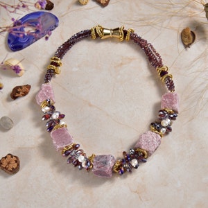 May include: A pink and purple gemstone necklace with gold accents. The necklace features a variety of beads, including small purple beads, white pearls, and larger pink gemstone beads.