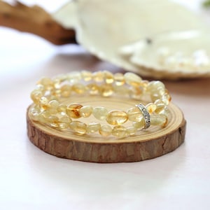 May include: Two yellow-orange gemstone bracelets with a silver accent ring are displayed on a small, round wooden slice. The bracelets are made of small, rounded stones. The background is blurred, with a white and light pink color scheme.