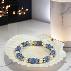 May include: A blue and white beaded bracelet with gold accents and white pearls. The bracelet is displayed on a white seashell.