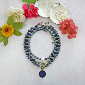 May include: A layered necklace with blue and green gemstone beads and a large blue pendant. The necklace is on a white background with flowers.