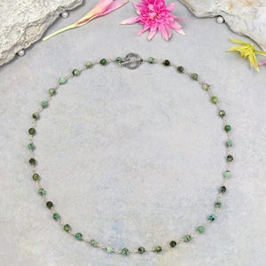 May include: A delicate necklace featuring small, round, turquoise-colored beads strung on a silver chain. The necklace has a decorative silver clasp and is displayed on a light gray surface with floral accents.