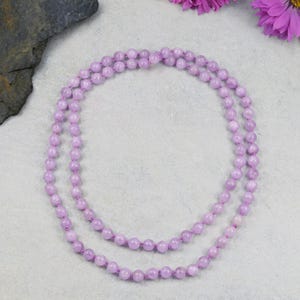 May include: A long, double-strand necklace composed of small, round, light purple beads. The beads are strung on a matching purple cord. The necklace is displayed on a light gray surface, with purple flowers in the background.