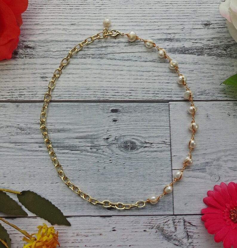May include: A gold chain necklace with a pearl and gold chain design. The necklace is on a white wooden background.