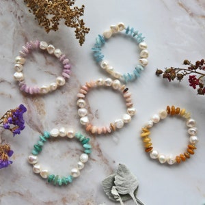 May include: Five pearl bracelets with different colored stones. The bracelets are on a white marble surface. The stones are pink, blue, peach, green, and yellow.