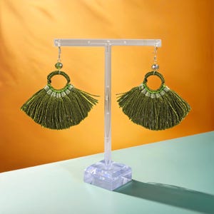 Könnte beinhalten: Ein Paar grüne Quastenohrringe mit einem silbernen Ring und einer kleinen grünen Perle. Die Ohrringe werden auf einem durchsichtigen Ständer vor einem gelben und blauen Hintergrund präsentiert.
