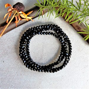 Puede incluir: Un collar de cuentas de cristal negro con cuentas facetadas. El collar está enrollado sobre una superficie gris.