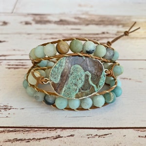 May include: A multi-strand bracelet featuring a large, oval-shaped, green and brown agate stone charm. The bracelet is crafted with brown leather cord and light green and brown beads.