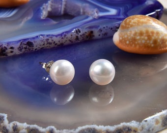Pode incluir: Um par de brincos de pérola branca com pinos de prata. Os brincos são redondos e têm uma superfície lustrosa. Os brincos são exibidos em uma pedra de ágata azul e cinza, com conchas no fundo.