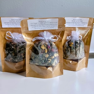 May include: Three bath soak products in clear bags with brown paper backing. Each bag contains dried herbs and flowers, tied with a white ribbon. The labels read "Soothe Bath Soak", "Cottage Flower Garden Bath Soak", and "Relax Bath Soak".