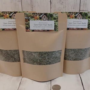 May include: Three brown paper bags with a clear window, each labeled "Sore Feet Foot Bath." The bags contain a green, dried herb mixture. The labels have a small image of a birdhouse and foliage.