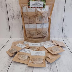 May include: A collection of tea bags in small brown paper packets, some tied with twine, and arranged on a light wood surface. The main package reads "Get Well Soon Tea Bags". The tea bags are labeled with the brand "Wood House Farm".