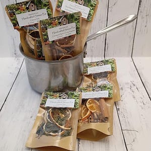 May include: A stainless steel saucepan containing three pouches of Handcrafted Simmer Pot mix, with two additional pouches placed in front. Each clear plastic pouch features a label with the Wood House Farm logo, scent name, and weight in grams.