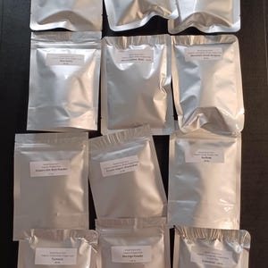 May include: Twelve silver pouches containing various organic powders, including marshmallow root, turmeric, and moringa powder. Each pouch is labeled with the name of the powder and the weight, 0.5 oz.