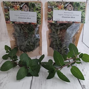 May include: Two clear pouches of air-dried chocolate mint, labeled "Wood House Farm." The pouches are filled with dried herbs and sit on a white wooden surface. Fresh green mint sprigs are in the foreground.