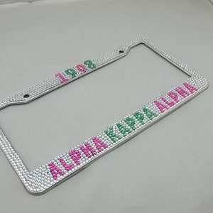 May include: A rectangular license plate frame covered in clear rhinestones. The text 1908 is spelled out in pink and green rhinestones at the top. The text ALPHA KAPPA ALPHA is spelled out in pink and green rhinestones along the bottom edge.