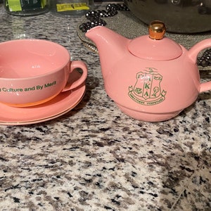 May include: Pink tea set with a teapot and a cup and saucer. The teapot has a gold lid and a green emblem with the letters AKA. The cup and saucer have the words "By Culture and By Merit" printed on them.