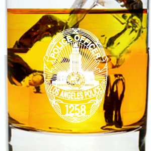 May include: A clear glass tumbler filled with a yellow liquid and ice cubes. The glass features a white Los Angeles Police Officer badge with the words "Police Officer" and "Los Angeles Police" and the number "1258" on it.