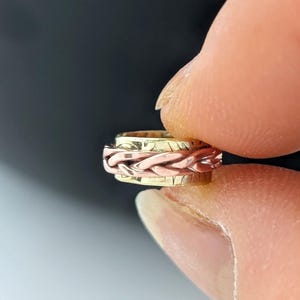 Può includere: Un anello in metallo multicolore caratterizzato da una fascia centrale intrecciata con finitura color rame, affiancata da due fasce lisce color oro. Il design incorpora un meccanismo rotante in cui la sezione centrale ruota indipendentemente.