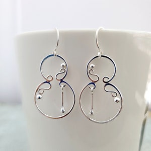 Puede incluir: Un par de pendientes de aro de plata con un diseño abstracto único. Los pendientes tienen un círculo con un círculo más pequeño en el interior y una cuenta colgante en la parte inferior.