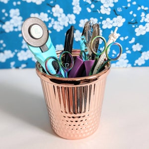 May include: A rose gold metal cup filled with various craft tools. The tools include a teal rotary cutter, scissors with rainbow-colored handles, a black pen, and a purple tool. The background is a blue floral pattern.