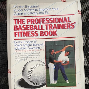 May include: A white book titled "THE PROFESSIONAL BASEBALL TRAINERS' FITNESS BOOK" with a baseball graphic and images of baseball trainers. The book cover has red and blue text and a baseball stitch design around the edges.