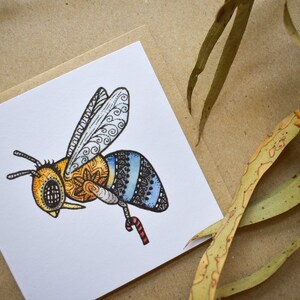 Könnte beinhalten: Quadratische Grußkarte mit detaillierter Illustration einer Biene mit komplizierten Mustern in Gelb, Orange, Blau und Schwarz. Die Biene wird im Flug dargestellt, mit einer Zuckerstange. Die Karte befindet sich auf einem neutralen Hintergrund mit Eukalyptusblättern.