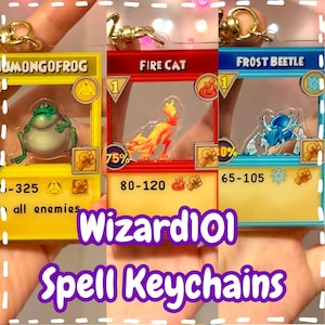 Może przedstawiać: Trzy breloczki do zaklęć Wizard101. Breloczki przedstawiają kolorowe wzory postaci z gry: zieloną żabę, ognistego kota i niebieskiego chrząszcza. Każdy breloczek ma złote zapięcie i zawiera statystyki gry. Widoczny jest tekst "Wizard101 Spell Keychains".