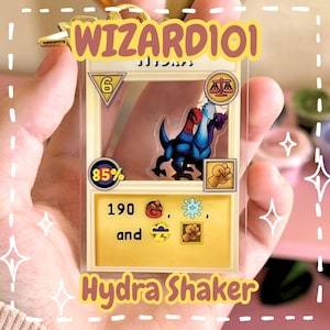 Peut inclure: Un porte-clés shaker en acrylique transparent illustrant une créature Hydra de Wizard101. Le design de la carte présente un fond jaune, un chiffre 6 dans un triangle, une icône de précision 85%, un symbole d'équilibre, un poing et des symboles de dégâts feu, glace, tempête et physiques.