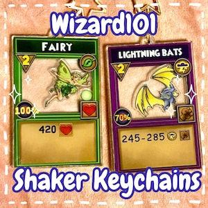 May include: Two acrylic shaker keychains featuring Wizard101 card designs. One shows a green Fairy with the text "FAIRY" and the other shows "LIGHTNING BATS". The keychains have a gold border and the text "Shaker Keychains" is at the bottom.