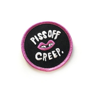 Op de afbeelding: Een ronde, geborduurde patch in zwart en roze met de tekst "Piss Off Creep." en een roze cartoonmond met tanden.