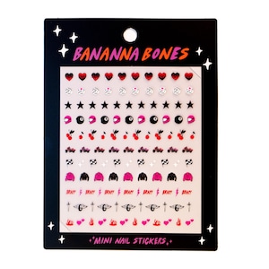 Puede incluir: Una hoja de pegatinas para uñas de color rosa y negro con varios diseños, incluyendo corazones, estrellas, cerezas, motocicletas y la palabra "BRAT!" La hoja está etiquetada como "BANANNA BONES +MINI NAIL STICKERS+"