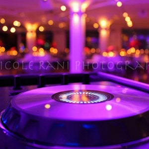 May include: A close-up of a DJ mixer with a silver record spinning on top. The mixer is lit with purple and blue lights, creating a vibrant and energetic atmosphere.