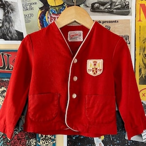 May include: Red vintage jacket with white piping and a crest on the left side. The jacket has a button closure and a pocket on the left side.