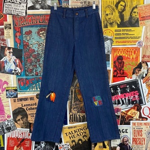 May include: Blue denim jeans with embroidered patches, including a flower on the left leg and the word "LOVE" on the right leg.