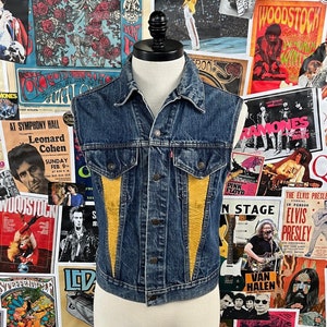 May include: A blue denim vest with yellow and brown leather accents. The vest has a zipper closure and two pockets on the front.