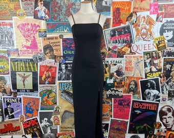 May include: A black maxi dress with a square neckline and thin straps. The dress is photographed against a wall covered in vintage concert posters.